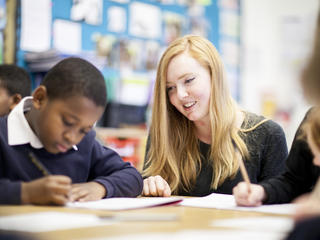 Female teacher smiling, looking at male student writing in notebook