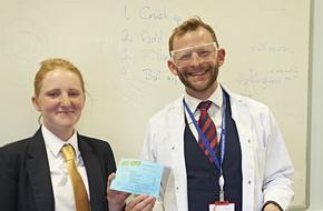 Headteacher in lab coat and goggles next to female student holding up a piece of paper