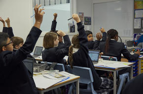 Image of pupils sat at desks in classroom, raising their hands