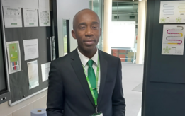 Junior, a SCITT trainee, stands in a classroom wearing a suit and green lanyard, smiling at the camera. Behind him, a blackboard with notices and a glass door with educational posters are visible.